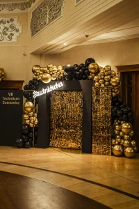 Personalizowana ścianka panelowa z nazwą szkoły i czarno-złotymi dekoracjami balonowymi, będąca częścią oprawy wizualnej eventu.