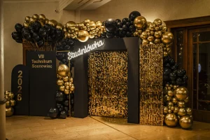 Złota ścianka cekinowa shimmer wall jako eleganckie tło do zdjęć na studniówkę, ozdobiona organiczną girlandą z czarno-złotych balonów