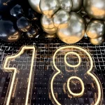 Zbliżenie na złote balony chrome i czarne marki Gemar w stylu organicznym wokół neonowego napisu 18 na tle czarnych cekinów.