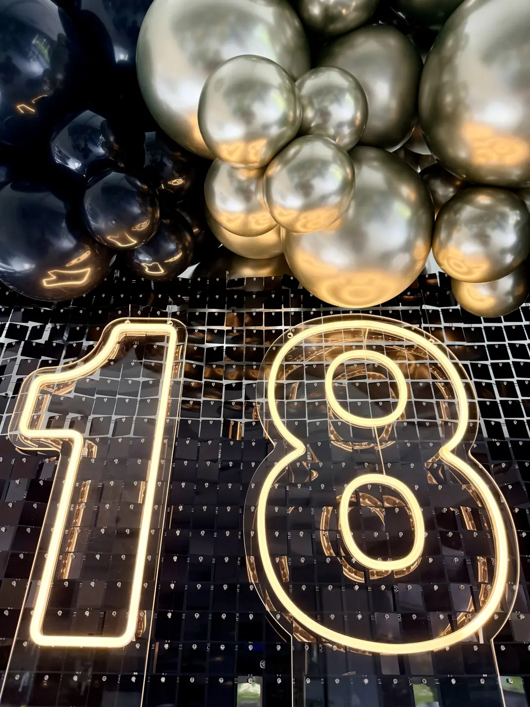 Zbliżenie na złote balony chrome i czarne marki Gemar w stylu organicznym wokół neonowego napisu 18 na tle czarnych cekinów.