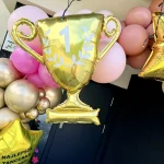Złote balony foliowe w kształcie pucharów oraz personalizowana gwiazda na szczycie dekoracji balonowej.